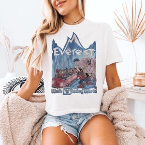 May include: White t-shirt featuring a graphic of Mickey and friends on a roller coaster with the text "Everest" and "Walt Disney World" in blue. The design includes a mountain range and a yeti. The shirt is a casual wear item.