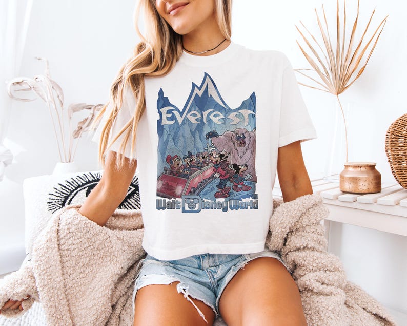 May include: White t-shirt featuring a graphic of Mickey and friends on a roller coaster with the text "Everest" and "Walt Disney World" in blue. The design includes a mountain range and a yeti. The shirt is a casual wear item.