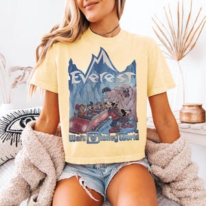 May include: Yellow t-shirt featuring a graphic of the Everest ride at Walt Disney World. The design includes the word "Everest" above a mountain scene with cartoon characters. The Walt Disney World logo is at the bottom.