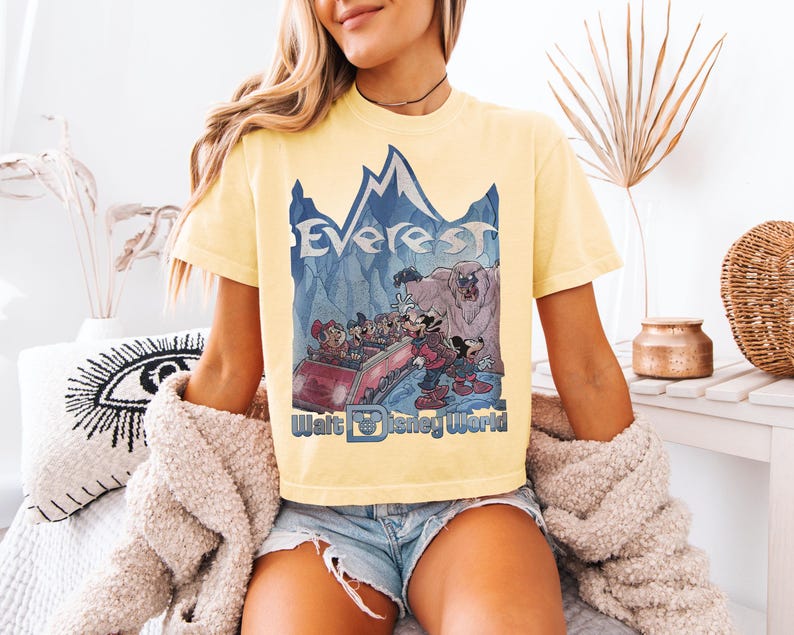 May include: Yellow t-shirt featuring a graphic of the Everest ride at Walt Disney World. The design includes the word "Everest" above a mountain scene with cartoon characters. The Walt Disney World logo is at the bottom.