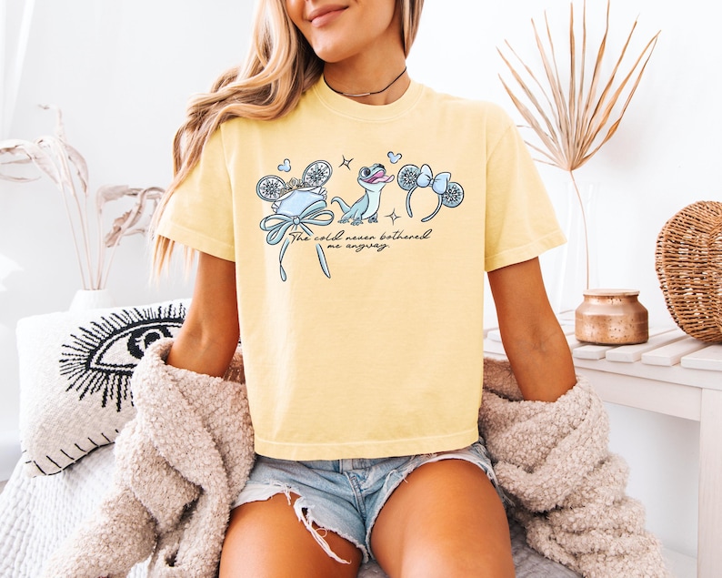 May include: Yellow t-shirt with a graphic featuring a blue cartoon lizard, a crown, and mouse ears. The text "The cold never bothered me anyway" is written in black script. The shirt is a light yellow color.
