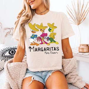 Op de afbeelding: Cr&egrave;mekleurig T-shirt met een cartoonontwerp van drie personages met sombrero's. De personages dragen blauwe, roze en gele outfits. De tekst "Margaritas Por Favor" staat eronder in het zwart.
