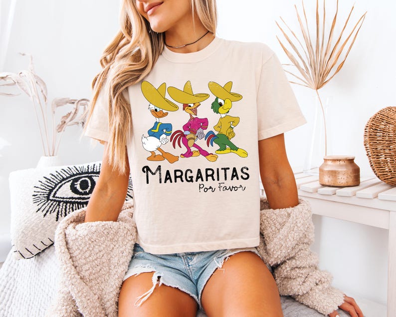 Op de afbeelding: Cr&egrave;mekleurig T-shirt met een cartoonontwerp van drie personages met sombrero's. De personages dragen blauwe, roze en gele outfits. De tekst "Margaritas Por Favor" staat eronder in het zwart.