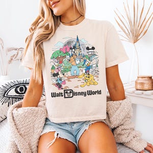May include: Cream-colored t-shirt featuring a colorful graphic of Disney characters and the text "Walt Disney World." The graphic includes Mickey and Minnie Mouse, Donald Duck, Goofy, and other characters in front of a castle.