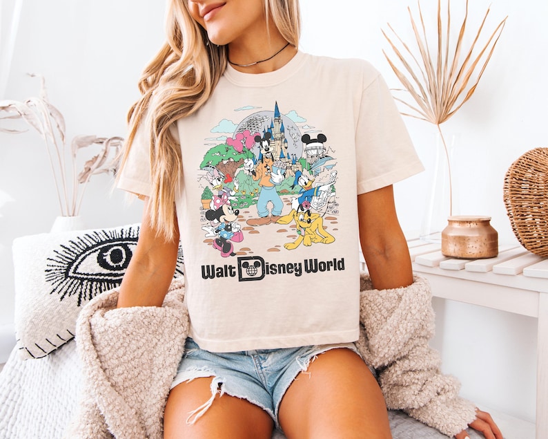 May include: Cream-colored t-shirt featuring a colorful graphic of Disney characters and the text "Walt Disney World." The graphic includes Mickey and Minnie Mouse, Donald Duck, Goofy, and other characters in front of a castle.