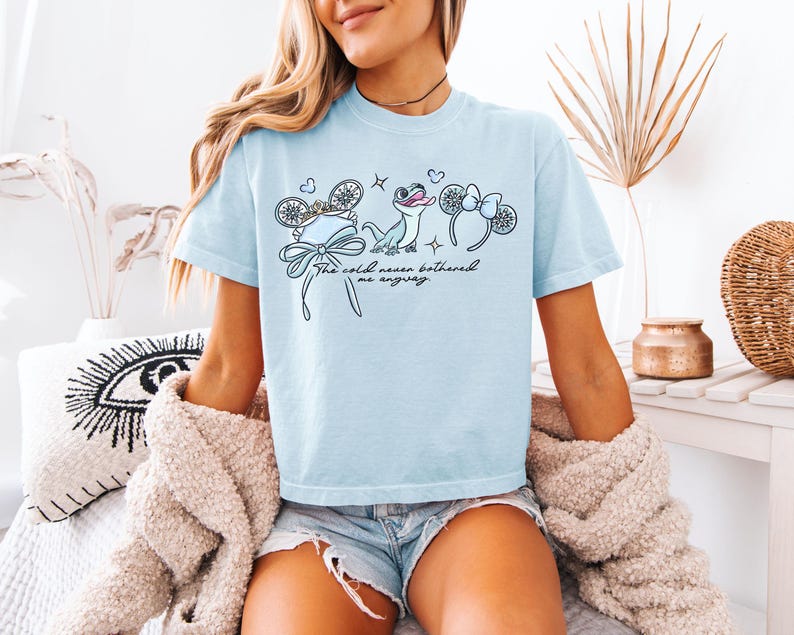 May include: Light blue t-shirt with a whimsical design featuring a bow, snowflakes, a frog, and mouse ears. The text "The cold never bothered me anyway" is written in a script font. The shirt is made of a soft material.