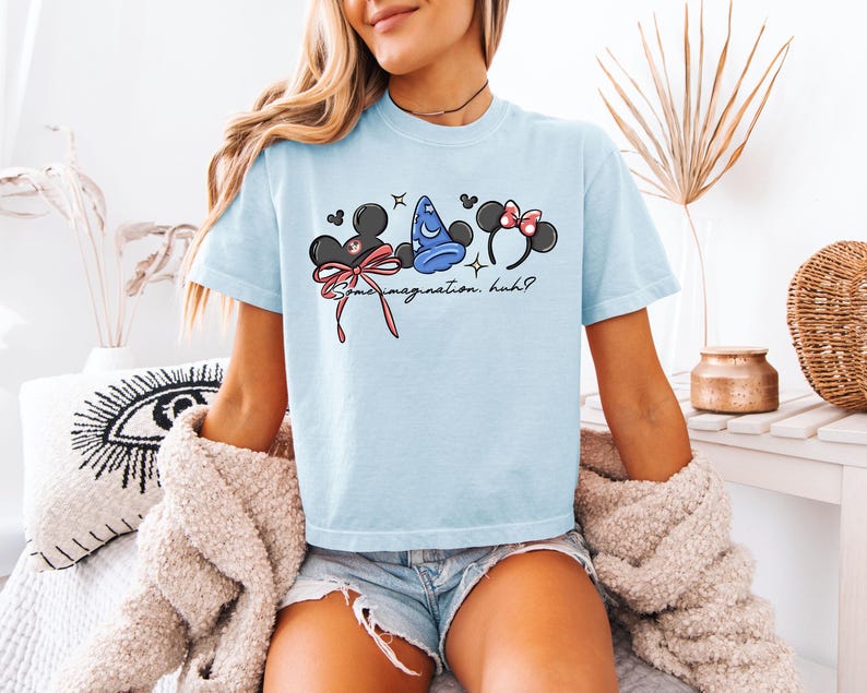 K&ouml;nnte beinhalten: Hellblaues T-Shirt mit einer Grafik von Mickey Mouse Ohren, Minnie Mouse Ohren und einem Zaubererhut. Der Text "Some imagination, huh?" ist darunter aufgedruckt. Das T-Shirt hat eine hellblaue Farbe.