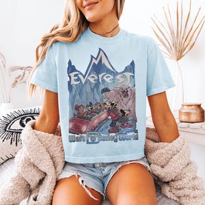 May include: Light blue t-shirt featuring a graphic of the Everest ride at Walt Disney World. The design includes the word "Everest" above a mountain scene with cartoon characters in a ride vehicle. The Walt Disney World logo is below.