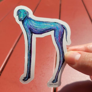 May include: A sticker of a blue and purple dog with long legs. The dog is standing on all fours and has a long tail. The sticker is on a red surface. The artist's name, "Ilana Napoli", is printed at the bottom of the sticker.