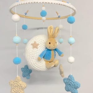 May include: A baby mobile with a crocheted moon and stars, featuring a plush Peter Rabbit toy sitting on the moon. The mobile has blue and white pom-poms and wooden beads.