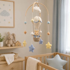 Puede incluir: Un móvil colgante para guardería que presenta un globo aerostático de ganchillo con un pequeño conejo de peluche en la cesta. El móvil incluye estrellas de ganchillo colgantes en amarillo y azul, junto con cuentas de madera en tonos pastel suaves.