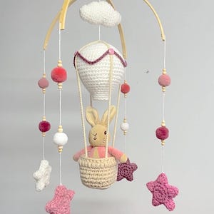 May include: A handcrafted baby mobile with a crochet hot air balloon and a bunny in a basket. The mobile includes hanging pink, white, and purple pom-poms, stars, and a cloud. The wooden arch and beads add a natural touch to this nursery decor.