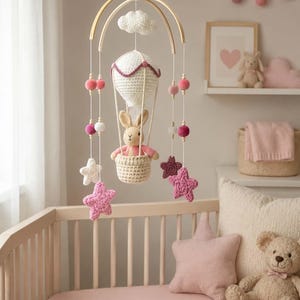 Könnte beinhalten: Ein hängendes Mobile mit einem gehäkelten weißen Heißluftballon, in dessen Korb ein kleiner beiger Hase sitzt. Das Mobile enthält hängende weiße und rosa gehäkelte Sterne, Holzperlen und rosa Pompons, die an einem gebogenen Holzrahmen befestigt sind.