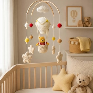 Peut inclure: Un mobile de chambre d'enfant composé d'une montgolfière crochetée avec une figurine d'ours jaune dans la nacelle, entourée d'étoiles crochetées, de nuages blancs et de boules en feutre suspendus à une structure en bois courbé.