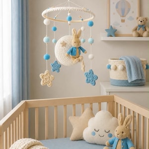 Puede incluir: Un móvil colgante para guardería con una luna de ganchillo, un conejo de peluche con chaqueta azul y pompones y estrellas azules y blancos. El móvil cuelga sobre una cuna de madera que contiene un conejo de peluche y un cojín de nube.