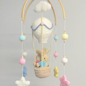 May include: A white crocheted hot air balloon with two plush bunny rabbits inside, hanging from a wooden mobile with a white crocheted cloud, stars, and colourful balls.