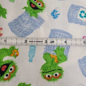 May include: White fabric with a repeating pattern of green Sesame Street character, Oscar the Grouch, holding a worm and standing in a blue recycling bin. The fabric also has a repeating pattern of blue recycling bins with a green recycling symbol. The fabric is measured with a ruler that reads "Made in Germany".
