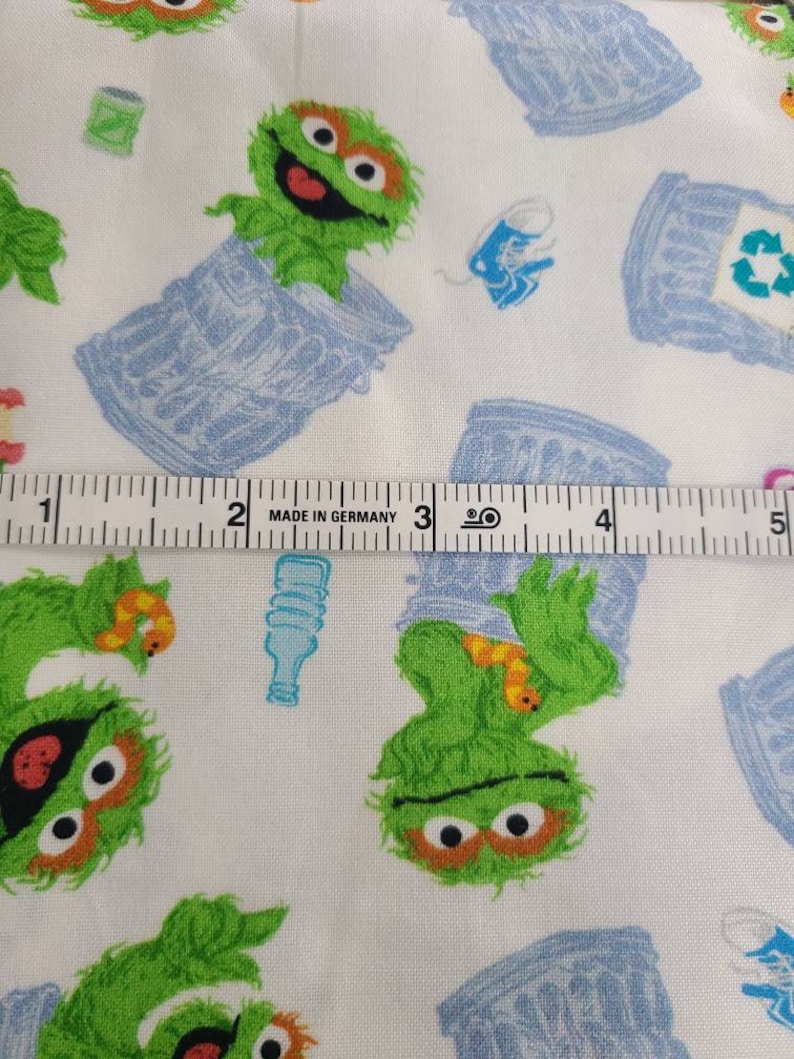 May include: White fabric with a repeating pattern of green Sesame Street character, Oscar the Grouch, holding a worm and standing in a blue recycling bin. The fabric also has a repeating pattern of blue recycling bins with a green recycling symbol. The fabric is measured with a ruler that reads "Made in Germany".