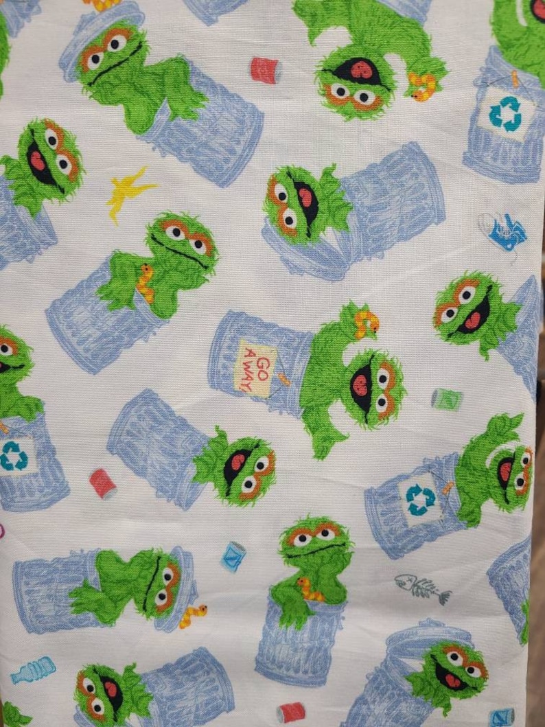 May include: White fabric with a repeating pattern of green Sesame Street character, Oscar the Grouch, holding a blue bin. The bin has a recycle symbol on it. The fabric also has a yellow lightning bolt and a red can.