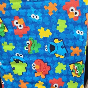May include: Blue fabric with a repeating pattern of Sesame Street characters, including Big Bird, Cookie Monster, and Elmo, in jigsaw piece shapes. The background is a solid blue with a subtle texture.