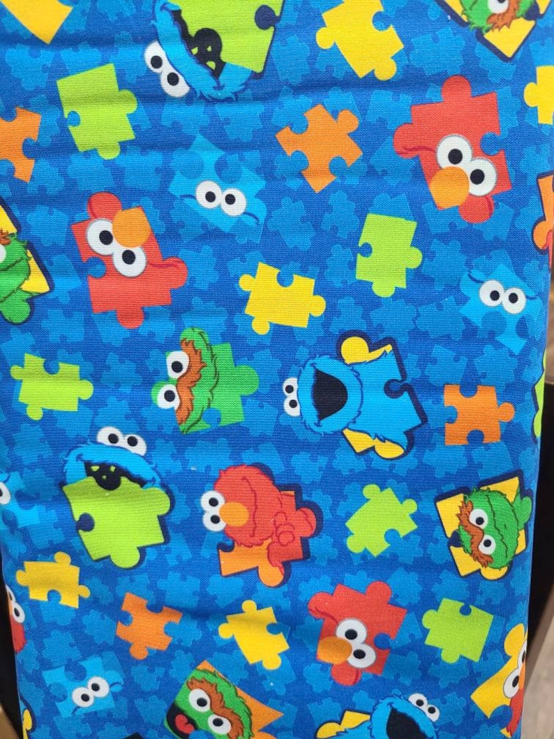 May include: Blue fabric with a repeating pattern of Sesame Street characters, including Big Bird, Cookie Monster, and Elmo, in jigsaw piece shapes. The background is a solid blue with a subtle texture.