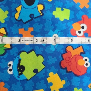 May include: Blue fabric with a repeating pattern of colourful puzzle pieces and Sesame Street characters, including Elmo and Cookie Monster. "Made in Germany" is printed on a ruler in the image.