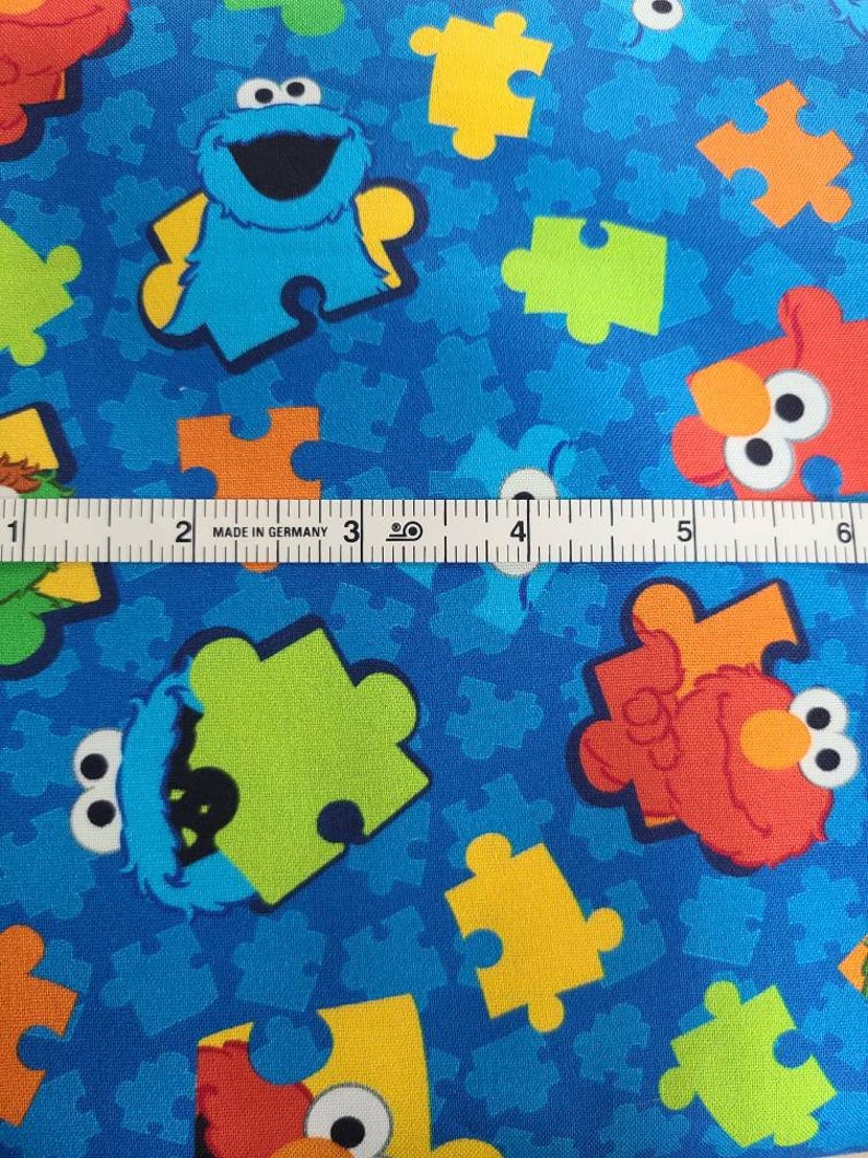 May include: Blue fabric with a repeating pattern of colourful puzzle pieces and Sesame Street characters, including Elmo and Cookie Monster. "Made in Germany" is printed on a ruler in the image.