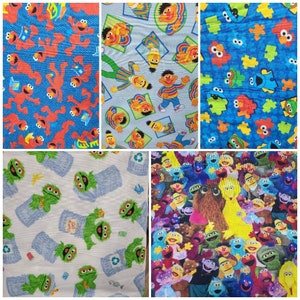 May include: Four different fabric swatches featuring Sesame Street characters. The first swatch is blue with red Sesame Street characters. The second swatch is blue with yellow Sesame Street characters. The third swatch is white with green Sesame Street characters. The fourth swatch is a colourful collage of Sesame Street characters.