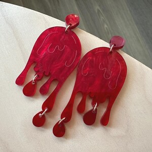 May include: A pair of red acrylic earrings with a dripping design. Each earring has a rounded top, a wavy interior design, and several dripping shapes that dangle from the bottom. The earrings are connected with silver-coloured metal rings.