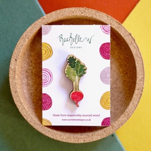 May include: A wooden pin shaped like a radish with green leaves. The pin is on a white card with the text "Rachelle W Designs" and "Made from responsibly sourced wood" and a website address. The card is on a cork coaster.
