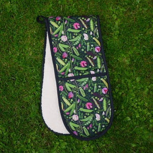 May include: Double oven glove featuring a dark background with a pattern of green pea pods and pink flowers. The interior is white. It has a black loop for hanging.