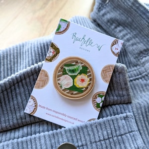 Puede incluir: Un pin de madera con una ilustración pintada a mano de un vaporizador de bambú lleno de tres albóndigas. El pin está hecho de madera de origen responsable y es un accesorio lindo y único. La tarjeta dice "Rachelle W Designs" y "Hecho de madera de origen responsable. www.rachellewdesigns.co.uk"