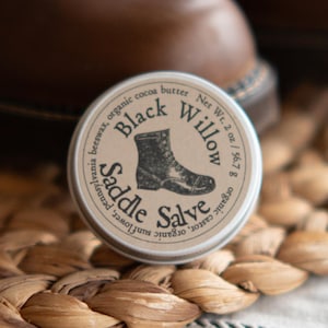 May include: A tin of Black Willow Saddle Salve with a boot illustration sits in front of a pair of brown leather boots. The salve tin is 2 oz / 56.7 g and contains organic ingredients. The boots are on a woven mat.