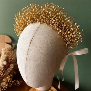 May include: A gold-coloured floral crown made of dried baby's breath flowers, featuring a dense arrangement of metallic gold-toned blossoms. The crown is secured with a thin, off-white satin ribbon tied into a bow at the back of the headpiece.