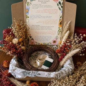 May include: A DIY hoop wreath kit in a cardboard box containing a brown grapevine wreath, a spool of green floral wire, a cream ribbon, and an assortment of dried flowers in shades of red, orange, and white, with an instructional guide.
