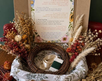 May include: A DIY hoop wreath kit in a cardboard box containing a brown grapevine wreath, a spool of green floral wire, a cream ribbon, and an assortment of dried flowers in shades of red, orange, and white, with an instructional guide.