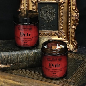 Könnte beinhalten: Zwei rot-schwarze beschriftete Kerzen mit dem Text "Dule" und "Well Witch Apothecary" auf alten Büchern. Die Kerzen befinden sich in Glasbehältern mit schwarzem Deckel. Die Bücher sind braun und haben ein goldenes Design auf dem Einband.