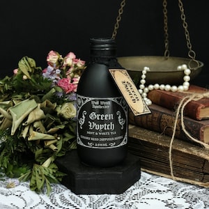 Könnte beinhalten: Eine schwarze Glasflasche mit einem Etikett, auf dem "Wolf Witch Apothecary Green Vvytch Mint & White Tea Luxury Reed Diffuser Refill 100 ML" steht. Die Flasche steht auf einem schwarzen sechseckigen Sockel. Die Flasche ist von getrockneten Blumen und einer antiken Waage umgeben.