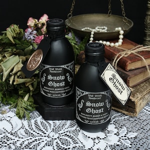 Könnte beinhalten: Zwei schwarze Glasflaschen mit schwarzen Etiketten und weißem Text, der "Snow Ghost" und "Wolf Wytch Apothecary" lautet. Die Flaschen stehen auf einer weißen Spitzentischdecke mit einem Stapel alter Bücher und einer Perlenkette auf einer Waage im Hintergrund.