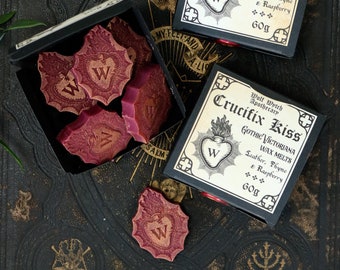Peut inclure: Deux boîtes noires avec des inscriptions dorées et un motif de cœur. Les boîtes contiennent des galets de cire sur le thème gothique victorien. Les galets de cire sont en forme de cœur et de couleur rouge. Le texte sur les boîtes indique "Crucifix Kiss Gothic Victoriana Wax Melts Leather, Thyme & Raspberry 60g".