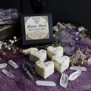 Può includere: Quattro tavolette da bagno a forma di cuore di colore bianco con lavanda e altre piante. Le tavolette sono su una superficie viola con cristalli di quarzo trasparenti e fiori secchi. L'etichetta recita "Mama Moon Herbal Bath Melts" con sfondo nero.