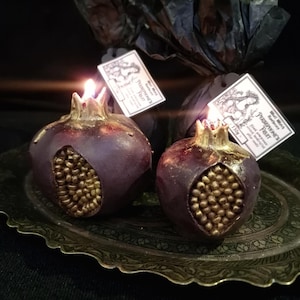 Puede incluir: Dos velas de color burdeos en forma de granada con detalles dorados sobre una bandeja de latón decorativa. Las velas están encendidas y tienen una etiqueta que dice "Tarta de granada" con una pequeña ilustración de una granada.