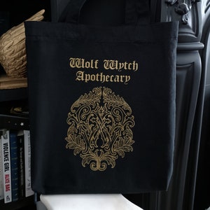 May include: Black tote bag with gold lettering that reads "Wolf Wytch Apothecary". A gold ornate design is centered on the bag.