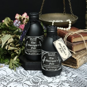 Könnte beinhalten: Zwei schwarze Glasflaschen mit schwarzen Etiketten und goldenen Schriftzügen. Die Etiketten lauten "Wolf Wytch Apothecary The Morrigan's Fate Blackcurrant & Licorice Luxury Reed Diffuser Refill 100 ml". Die Flaschen stehen auf einer schwarzen Oberfläche mit einer weißen Spitzentischdecke im Hintergrund.