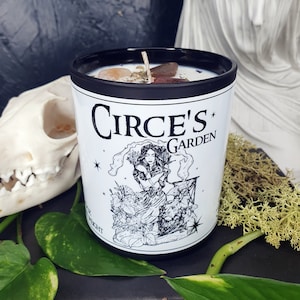 May include: A black candle jar with a white label that reads "Circe's Garden". The label features a black and white illustration of a woman with long hair standing in a garden. The candle jar is surrounded by green leaves and a white statue.