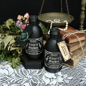 Könnte beinhalten: Zwei schwarze Glasflaschen mit Etiketten, auf denen "Wolf Wyteh Apothecary Circe's Garden Fresh Citrus & Herbs Luxury Reed Diffuser Refill 100 ML" steht. Die Flaschen stehen auf einer schwarzen Holzoberfläche mit einer weißen Spitzentischdecke darunter.
