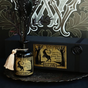 Könnte beinhalten: Ein schwarzes Apotheken-Schilfrohr-Diffusor-Set mit passender Box. Die Diffusorflasche hat eine beige Quaste und ein Etikett mit einer Ziegenillustration. Auf der Box steht "Wolf Wytch Apothecary" und "black phillip". Der Hintergrund ist ein dekoratives Muster.
