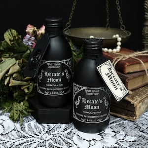 May include: Two black glass bottles with black labels that read "Wolf Wytch Apothecary Hecate's Moon Tobacco & Oak Luxury Reed Diffuser Refill 100ML". The bottles are sitting on a white lace tablecloth.