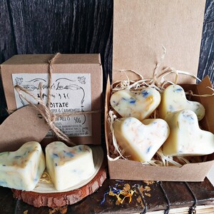 May include: Heart-shaped wax melts with blue and orange floral accents are displayed in a brown box and on a wooden slice. The box is labeled "Meditate" with Juniper & Chamomile. The melts are a creamy white color.