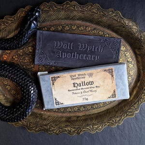 May include: A dark, ornate tray holds two bars of soap. One is a dark gray rectangle with "Wolf Wytch Apothecary" embossed on it. The other is a rectangular bar of soap in a gray box with a label that reads "Hallow" and "75g". A black snake decorates the tray.